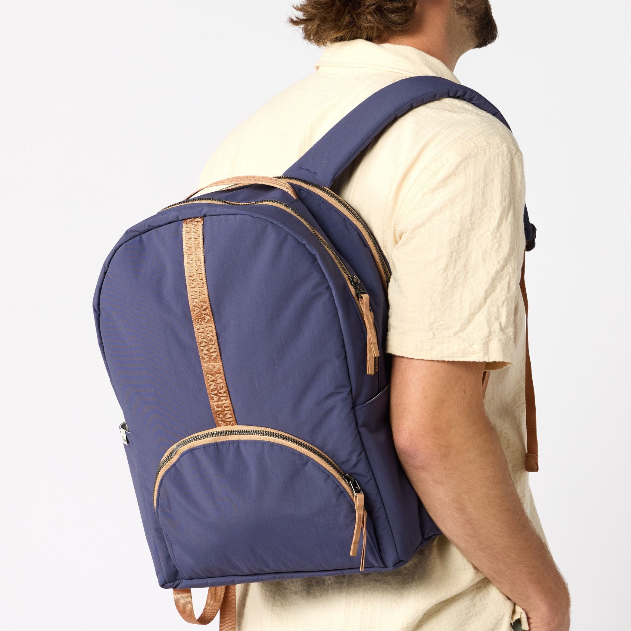 A man wearing a cream-colored shirt is shown from the back and side, carrying a navy blue backpack with tan leather-like trim and zipper pulls against a light gray studio background. #indigo_and_camel