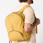 A man wearing a cream-colored shirt is shown from the back and side, carrying a mustard yellow backpack against a plain white background. #amber_yellow