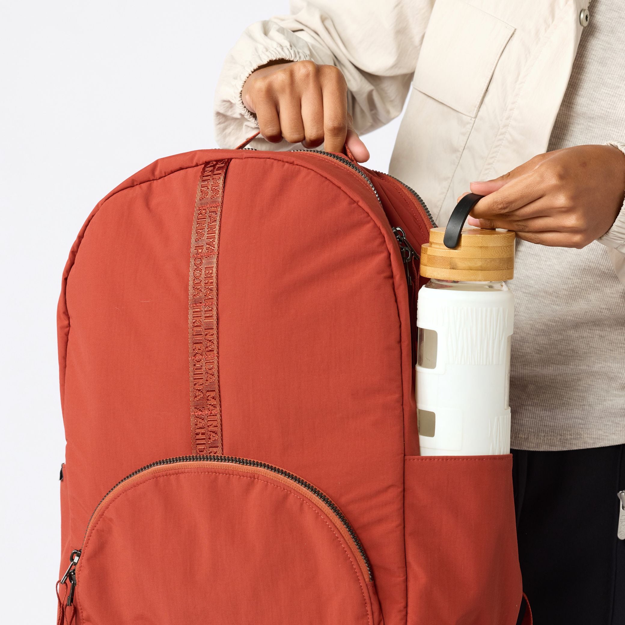 A terracotta orange backpack with a vertical textured stripe detail, shown with a person's hands loading a white water bottle into its side pocket. #rust_red