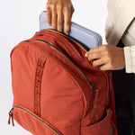 A close-up of a terracotta orange backpack being filled with a light blue laptop by a person wearing a cream shirt and black pants. The backpack has multiple zippered compartments and a vertical woven brand strap. #rust_red