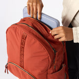 A close-up of a terracotta orange backpack being filled with a light blue laptop by a person wearing a cream shirt and black pants. The backpack has multiple zippered compartments and a vertical woven brand strap. #rust_red