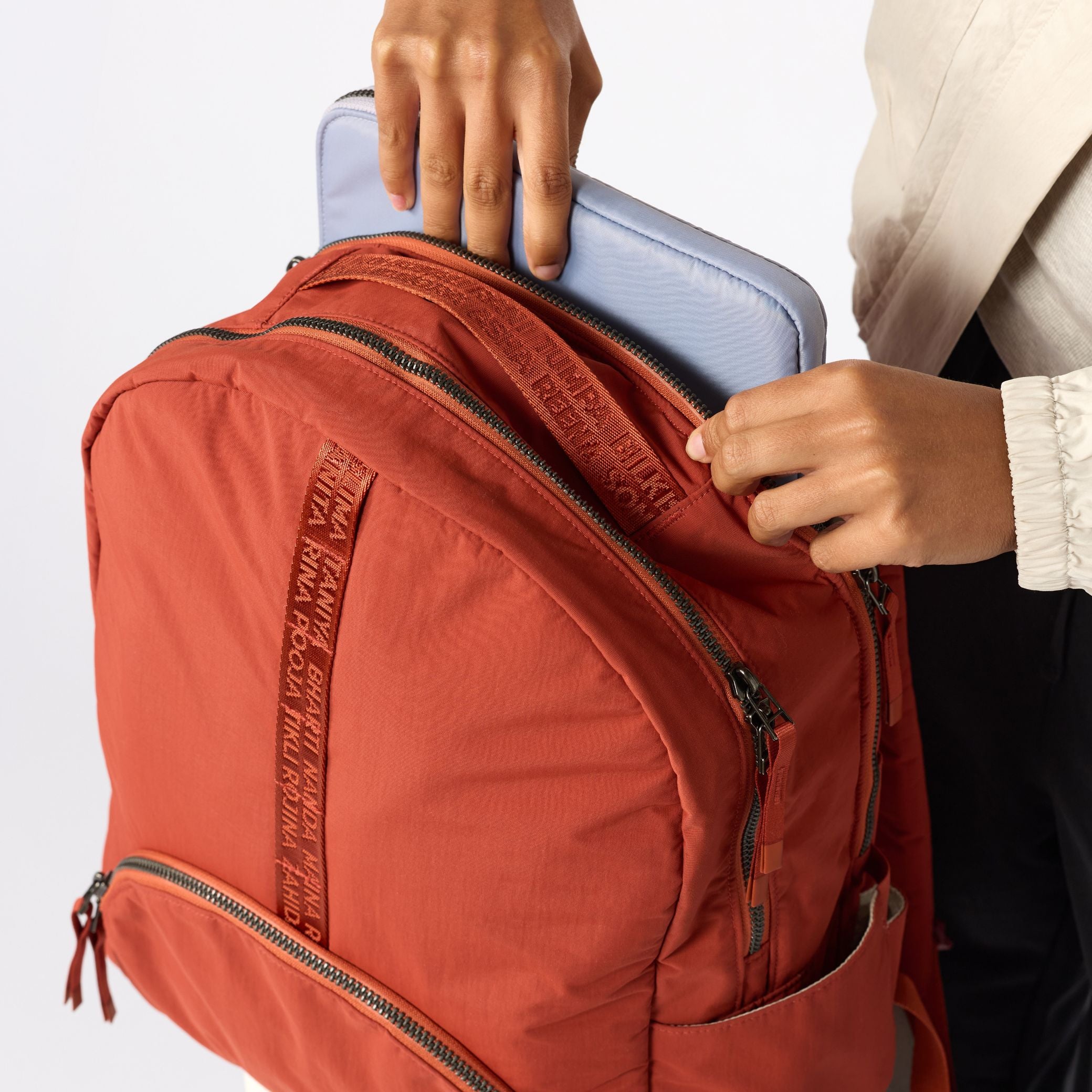 A close-up of a terracotta orange backpack being filled with a light blue laptop by a person wearing a cream shirt and black pants. The backpack has multiple zippered compartments and a vertical woven brand strap. #rust_red