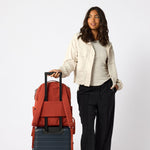 A woman stands next to a red backpack mounted on a dark blue rolling suitcase against a light gray studio background. #rust_red