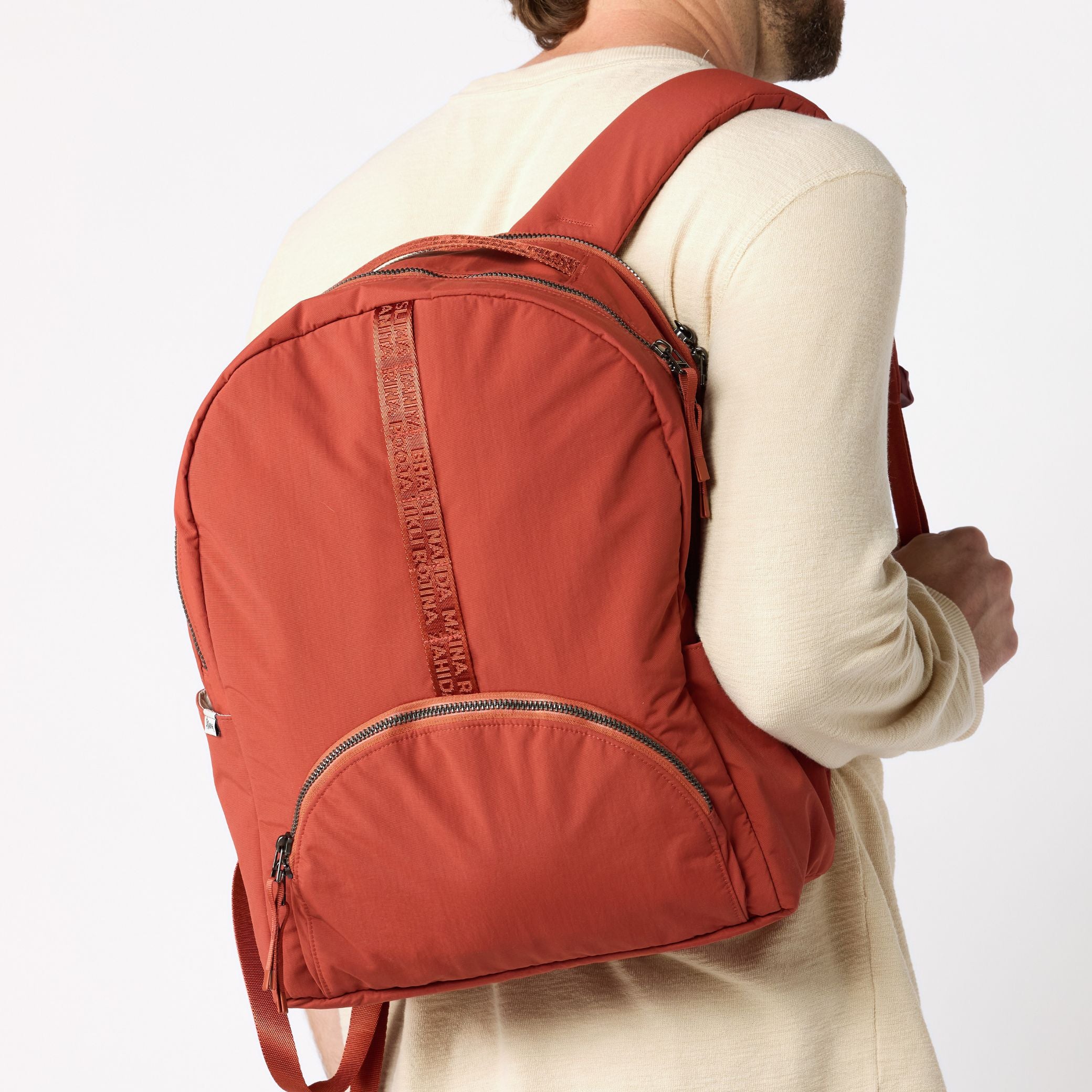 A man wearing a cream long-sleeve shirt is shown from the back, carrying a rust-orange nylon backpack with a front zippered pocket and a vertical textured strap detail. #rust_red