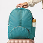 A teal blue backpack with a front zippered pocket and side bottle holder, shown with a hand holding its top handle and resting on top of a rose gold suitcase against a light gray  #solid_teal