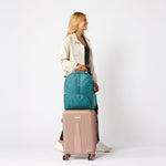 A woman stands in profile holding a teal backpack stacked on top of a pink rolling suitcase against a clean light gray studio background. #solid_teal