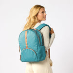A woman with blonde hair is shown from the back wearing a teal blue backpack with tan leather-like trim straps, against a plain white studio  #teal_and_camel