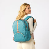 A woman with blonde hair is shown from the back wearing a teal blue backpack with tan leather-like trim straps, against a plain white studio  #teal_and_camel
