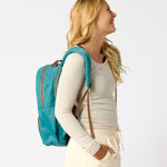 A woman in a cream ribbed long-sleeve top and matching drawstring pants is shown from the side, wearing a bright turquoise backpack with brown straps and zipper pulls against a plain white studio background. #teal_and_camel