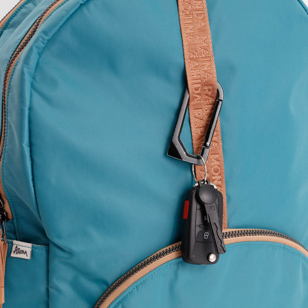 Close-up of a turquoise blue nylon backpack with a tan monogrammed shoulder strap attached to a black car key fob holder. #teal_and_camel