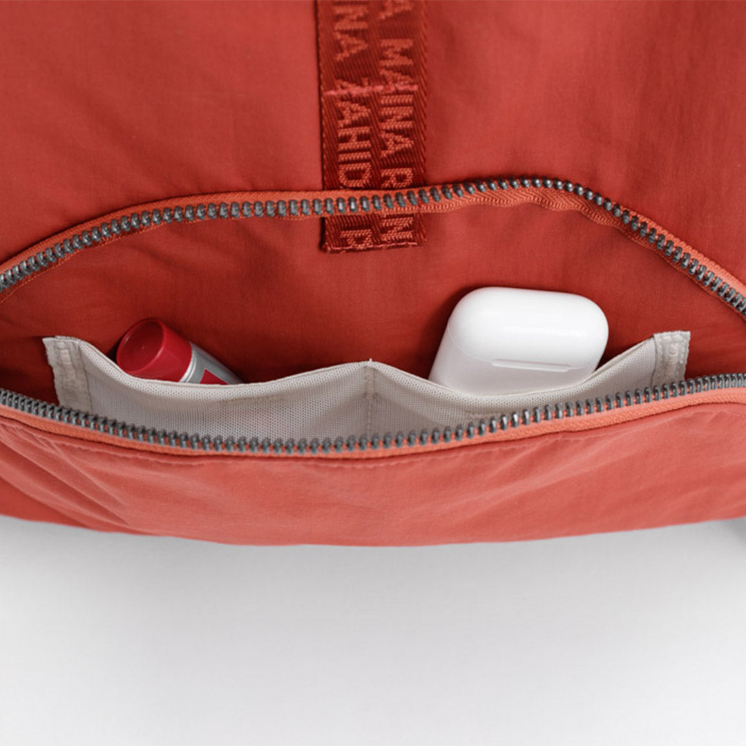 Close-up of a coral-orange zippered pouch or backpack interior showing a light gray organizer pocket with a white earbud case and a small red-and-white tube, plus a vertical branded fabric #rust_red