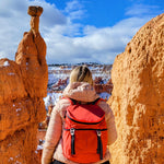 Person with a red sonu backpack standing between two large orange rock formations with a snowy landscape in the background. #poppy_red